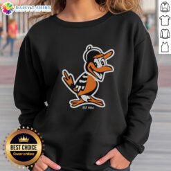 Hot Zay wearing a Baltimore Orioles mascot sweatshirt, celebrating team spirit since 1954 with vibrant flowers in the background.